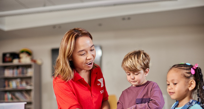 Preschool teacher with a preschool aged boy and girl sitting around a table. The teacher is pointing to the an item on the table,