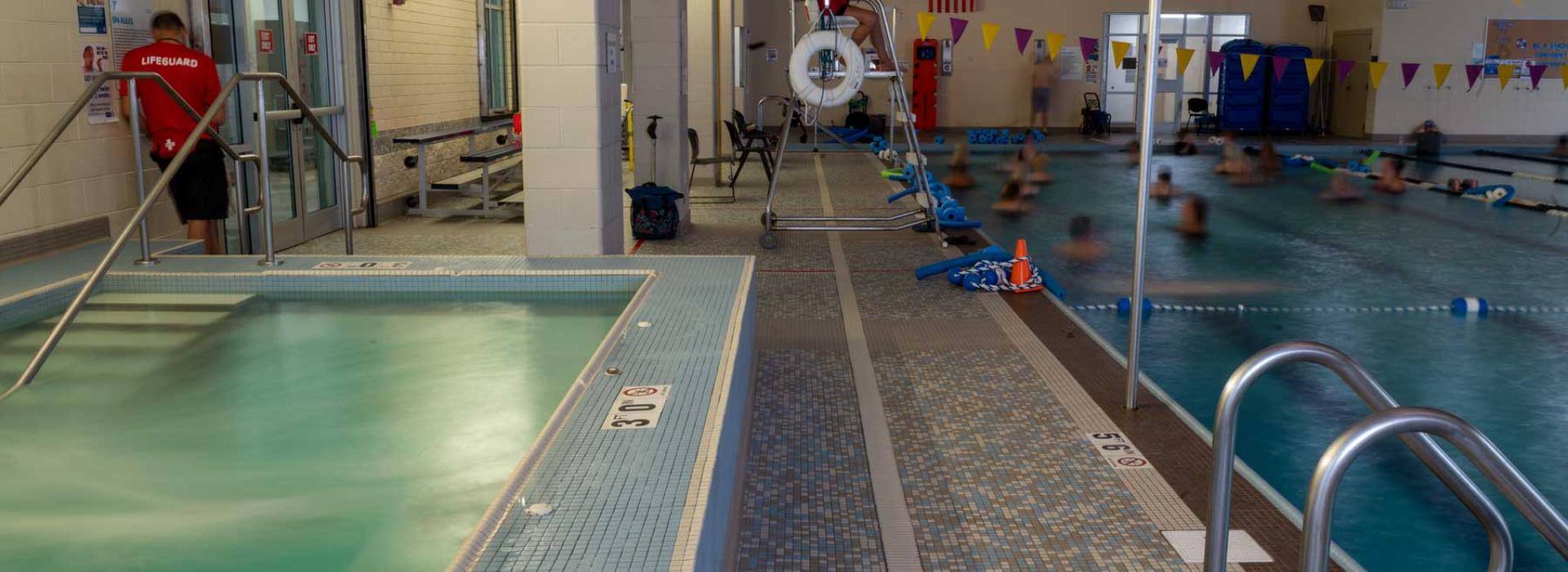 Image shows the North Royalton pool and hot tub. Hot tub on left side of image as a water-exercise class is pictured in the pool to the right. Pictured: 2 lifeguards overseeing swimming area.