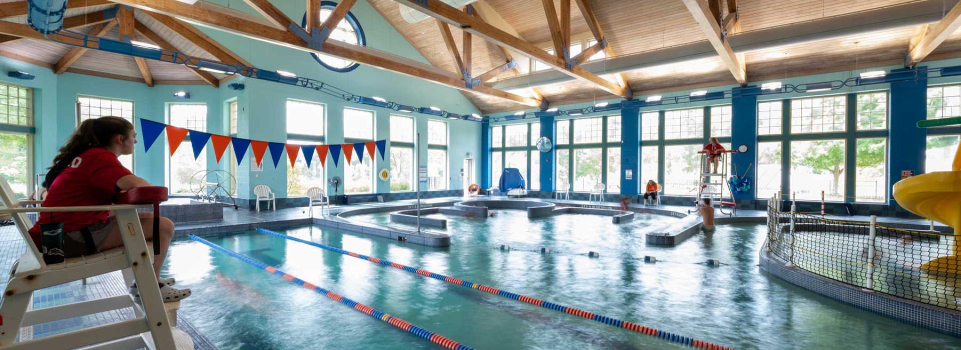 Image shows female lifeguard looking over pool area. Image of pool area includes 3 lanes for swimming, hot tub with &quot;lazy river&quot; around it, kids playing in pool, and jungle-gym water park slide.